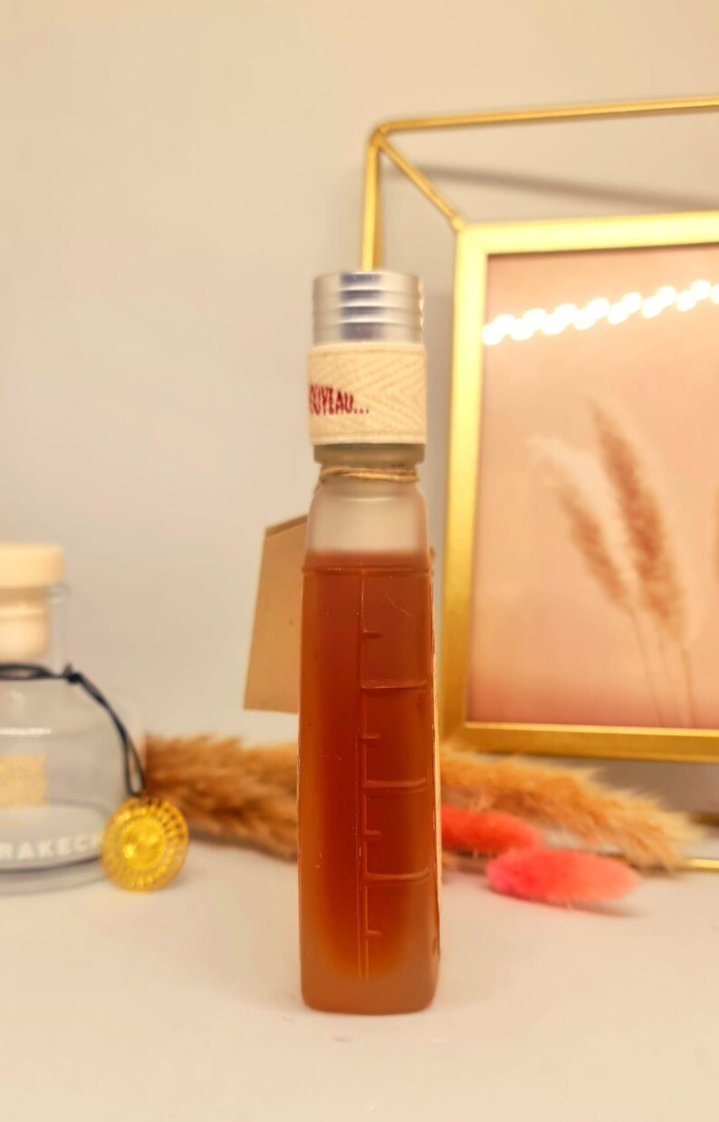 Pu&ograve; includere: Una bottiglia rettangolare trasparente riempita con un liquido marrone, con un tappo argentato e un'etichetta che recita "NUVEAU...". La bottiglia ha segni di misurazione sul lato e un piccolo cartellino. Lo sfondo include una cornice dorata ed elementi decorativi.
