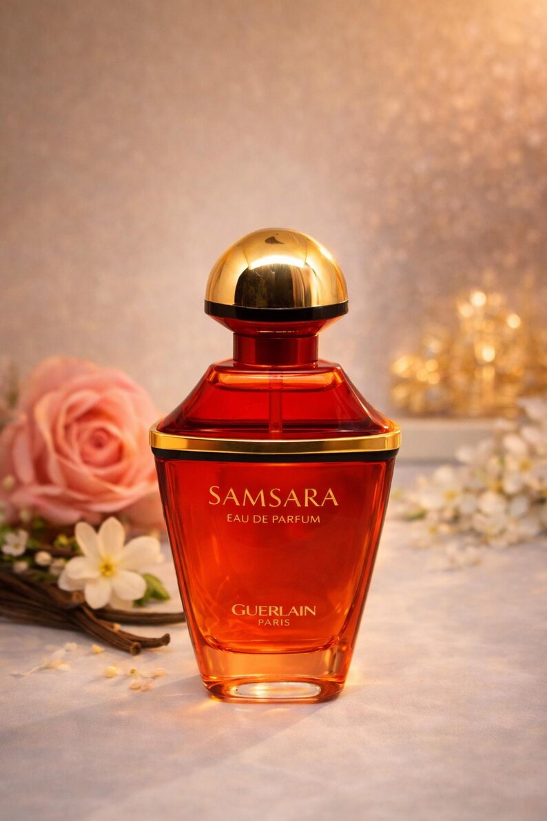 Może przedstawiać: Butelka perfum Samsara Eau de Parfum ze złotą nakrętką i czerwono-pomarańczowym korpusem. Butelka ma złote obramowanie i widoczne jest logo Guerlain Paris. R&oacute;żowa r&oacute;ża i białe kwiaty są w tle.