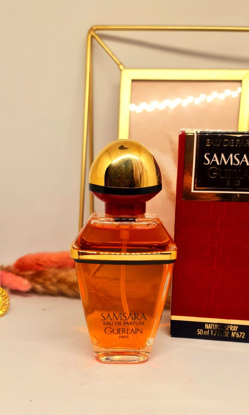 Może przedstawiać: Butelka perfum Samsara z złotą nakrętką i czerwonym pudełkiem. Butelka perfum ma przezroczysty, geometryczny kształt ze złotą obw&oacute;dką. Pudełko jest czerwone ze złotymi literami. Zawiera 50 ml perfum.