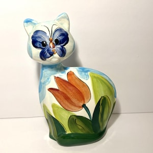 May include: A ceramic cat figurine with a hand-painted design. The cat's face features a blue butterfly, and its body is decorated with a large orange tulip and green leaves. The figurine has a white base with blue accents.