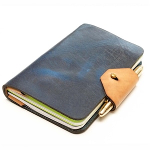 May include: A dark blue leather journal with a textured finish, featuring a light tan leather strap closure secured by a brass stud. A gold-toned metal pen is tucked into the strap, and the edges of the white paper pages are visible.