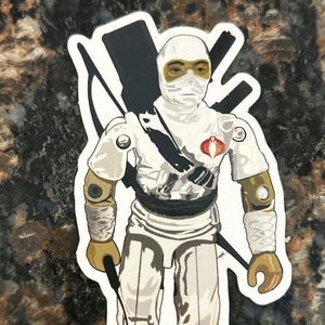 May include: A sticker featuring a white-clad action figure with a masked face and gold accents. The figure has a black belt, a red emblem, and carries a black weapon. The sticker is a collectible item.