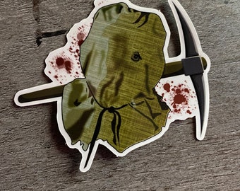 May include: A sticker of a green and brown burlap sack head with a pickaxe through it. The head has blood splatters on it.
