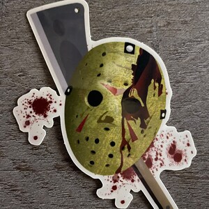 May include: A sticker featuring a white, green, and red hockey mask with blood splatters and a butcher knife. The mask has a black eye hole and red stripes.
