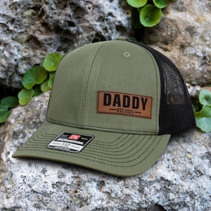 May include: An olive green trucker hat with a black mesh back. A brown leather patch on the front reads "DADDY - EST.2025". The hat has a curved brim and a Richardson 112 tag.