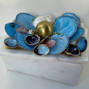 May include: A white marble box with a blue and gold seashell design. The box is decorated with a variety of seashells, including blue, purple, and pink shells. The box is a unique and decorative piece for any home.
