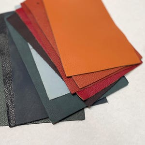 May include: A fan of leather swatches in various colors, including orange, red, brown, and green. The swatches are arranged in a layered pattern, showcasing the different textures and shades of the material. The leather appears to be smooth and textured.