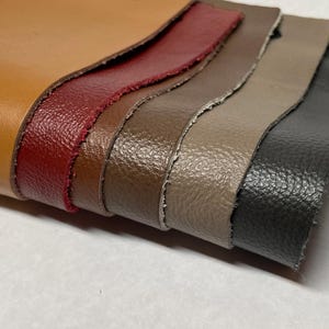 May include: A stack of leather swatches in various colors, including tan, burgundy, brown, taupe, and charcoal gray. The edges of the leather are rough-cut, revealing the texture of the material. The swatches are layered, showcasing the different shades and textures.