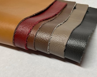 May include: A stack of leather swatches in various colors, including tan, burgundy, brown, taupe, and charcoal gray. The edges of the leather are rough-cut, revealing the texture of the material. The swatches are layered, showcasing the different shades and textures.
