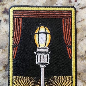 May include: An embroidered patch featuring a white spotlight with a yellow globe on a black background. The spotlight is on a stage with red curtains and a yellow and black striped floor.
