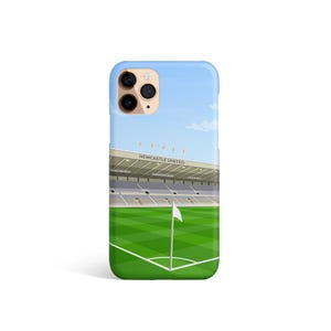 Może przedstawiać: Etui na telefon z wzorem stadionu piłkarskiego z zielonym boiskiem i białą flagą. Na stadionie widnieje napis "NEWCASTLE UNITED". Etui ma czarny wystający aparat z trzema obiektywami.