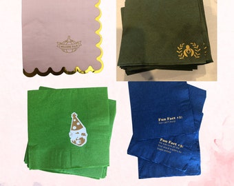 May include: Four stacks of custom napkins with different designs. The first stack is pink with gold scalloped edges and the text "WELCOME ELLIE" with a pumpkin graphic. The second stack is dark green with a gold graphic of a woman holding a baby. The third stack is green with a gold graphic of a man wearing a party hat. The fourth stack is blue with the text "Fun Fact #1: Kev can't burp" and "Fun Fact #3: He was one of the first stock traders to trade online".