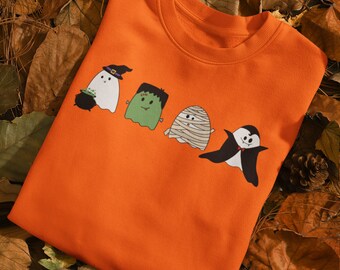 May include: Orange sweatshirt with a graphic print of four Halloween characters: a ghost with a witch hat, a green monster, a mummy, and a vampire ghost.