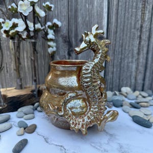 May include: A ceramic vase with a seahorse design in shades of gold and brown. The seahorse is sculpted on the side of the vase, adding a unique decorative element. The vase is likely used for displaying flowers or as a decorative piece.