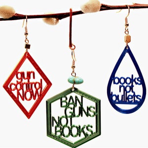 May include: Three pairs of earrings with different shapes and colors. The first pair is red and diamond-shaped with the text "gun control now". The second pair is green and hexagonal with the text "ban guns not books". The third pair is blue and teardrop-shaped with the text "books not bullets".