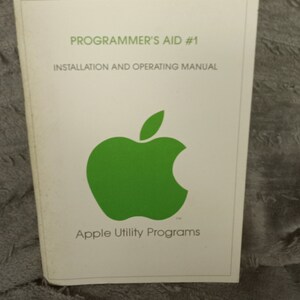 May include: A white cover for a Programmer's Aid #1 Installation and Operating Manual for Apple Utility Programs. The cover features a green Apple logo.