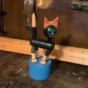 May include: A black wooden cat puppet with red ears and green eyes. The cat is standing on a blue wooden base.