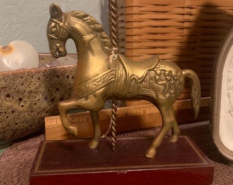 May include: A vintage brass carousel horse figurine. The horse is mounted on a dark red wooden base. The horse has intricate detailing and a gold finish. The carousel horse is a decorative item.