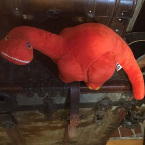 May include: A bright orange plush dinosaur toy with a white mouth and purple eyes. The dinosaur is sitting on top of a vintage wooden trunk with metal accents.