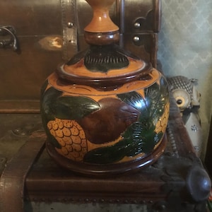 May include: A wooden decorative jar with a lid and a knob on top. The jar is painted with green leaves and yellow flowers. The jar is sitting on top of a wooden chest.