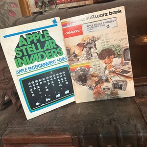Peut inclure: Une cartouche de jeu vintage Apple II pour Apple Stellar Invaders, faisant partie de la série Apple Entertainment Series. La cartouche est dans son emballage d'origine, qui présente une illustration colorée de personnes utilisant un ordinateur Apple II. L'emballage comprend également le texte "Apple Stellar Invaders" et "Apple Entertainment Series".