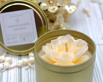 May include: A gold tin candle with a white flower-shaped wax design. The label on the lid reads "Indian Jasmine (Mogra)".