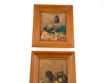 Puede incluir: Dos pinturas enmarcadas de naturalezas muertas. La pintura superior presenta una linterna, libros y una pequeña caja. La pintura inferior muestra uvas, un libro y un candelabro. Ambas pinturas están en marcos de madera.