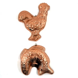 May include: Two copper-colored molds: a rooster and a fish. The rooster mold is upright, with detailed feathers and a comb. The fish mold is horizontal, with scales and fins. Both molds have a loop for hanging, and are set against a white background.