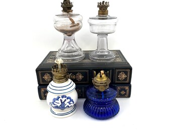 May include: Four antique oil lamps. Two clear glass lamps with brass accents and two ceramic lamps, one blue and white, the other a deep blue glass. The lamps are displayed on a stack of dark books.