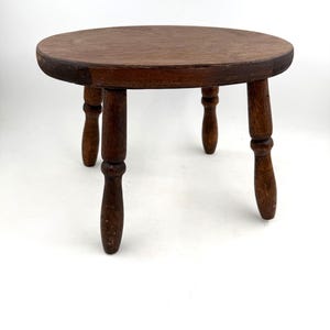 May include: A small, round, dark brown wooden stool featuring a smooth, circular top surface supported by four turned legs. The wood grain is visible across the top, and the legs have a distinct, rounded, spindle-like shape with a polished finish.
