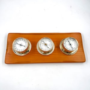 May include: A wooden weather station featuring three round instruments with silver-colored bezels and white faces. The instruments are mounted on a rectangular, light brown wooden base. The dials have black markings and needles.