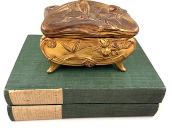 Puede incluir: Una pequeña joyero antiguo de color dorado con detalles florales en relieve en la tapa y el cuerpo. La caja está sobre tres pequeños pies y sobre dos libros de tapa dura verdes. La caja mide aproximadamente 10 cm de largo.
