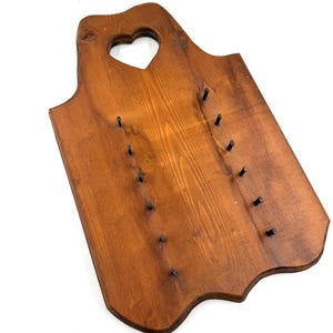 May include: A rectangular brown wooden wall-mounted display board featuring a heart-shaped cutout at the top center. The board has twelve small black metal pegs arranged in two vertical rows of six, designed for hanging items on the surface.