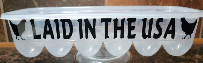 May include: A clear plastic egg carton with a black silhouette of a chicken on each end and the text "LAID IN THE USA" printed in black.