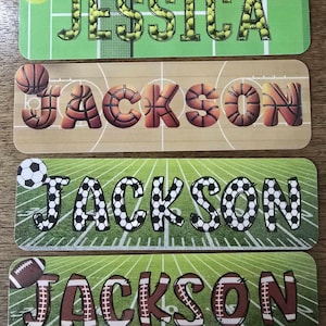 May include: Four personalized name tags with sports themes. The top tag says "JESSICA" with tennis ball-filled letters on a green tennis court background. The other tags say "JACKSON" with basketball, soccer, and football-themed letters.
