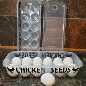 May include: A clear plastic egg carton with a black silhouette of a chicken and the text "Chicken Seeds" on the front. The carton is filled with white eggs.