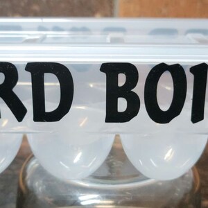 May include: A clear plastic egg carton with a black silhouette of a chicken on each end and the text "HARD BOILED" in black letters.