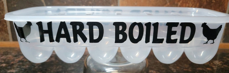 May include: A clear plastic egg carton with a black silhouette of a chicken on each end and the text "HARD BOILED" in black letters.