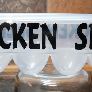 May include: A clear plastic egg carton with a black silhouette of a chicken on each end and the text "CHICKEN SEEDS" in black lettering.