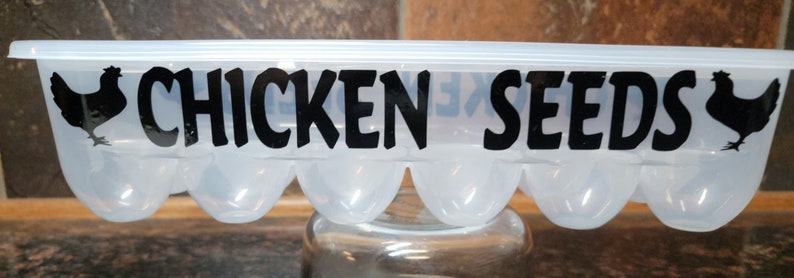 May include: A clear plastic egg carton with a black silhouette of a chicken on each end and the text "CHICKEN SEEDS" in black lettering.