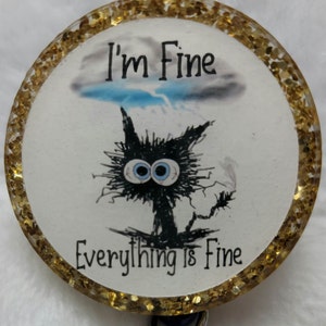 May include: A round, gold glitter-rimmed badge with a black cat with blue eyes and a storm cloud above it. The text "I'm Fine" is above the cat and "Everything is Fine" is below the cat.