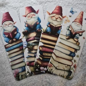May include: Four whimsical bookmarks featuring illustrations of gnomes reading books. Each gnome wears a red hat and blue outfit, sitting atop a stack of books. The bookmarks have a white background with floral and butterfly accents.