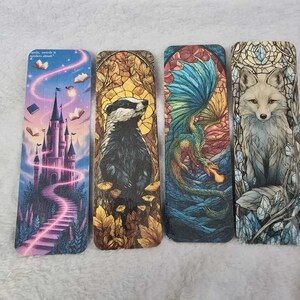 May include: Four rectangular bookmarks with fantasy-themed illustrations. One features a castle with a winding path, another a badger, a dragon, and a fox. The dragon has blue wings and the fox is surrounded by crystals. The first bookmark has the text "spells, words & spoilers ahead."