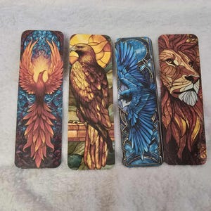 May include: Four rectangular bookmarks with animal designs. The bookmarks feature a phoenix, an eagle, a blue bird, and a lion. The phoenix is red and orange, the eagle is brown and yellow, the blue bird is blue, and the lion is brown and orange.