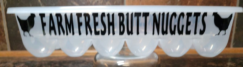 May include: White plastic egg carton with black chicken silhouettes and the text "FARM FRESH BUTT NUGGETS".