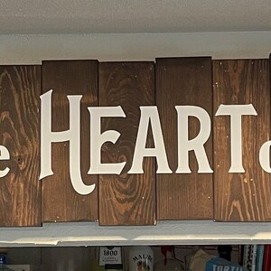 Puede incluir: Un letrero de madera con las palabras "Kitchen The HEART of the home" pintadas en blanco. El letrero está hecho de tablones de madera individuales.