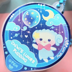 Peut inclure: Un charme bleu et blanc avec un mouton de dessin animé mignon tenant un ballon. Le charme dit "Chiwi's Lunar Pudding" et "Make with love".