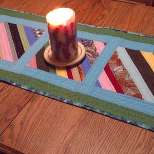Op de afbeelding: Handgemaakte tafelloper met een patchwork ontwerp met diagonale strepen in verschillende kleuren. Een brandende kaars staat op een houten onderzetter in het midden. De tafelloper ligt op een houten tafel. Ongeveer 76 cm lang.