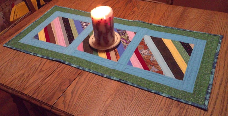 Op de afbeelding: Handgemaakte tafelloper met een patchwork ontwerp met diagonale strepen in verschillende kleuren. Een brandende kaars staat op een houten onderzetter in het midden. De tafelloper ligt op een houten tafel. Ongeveer 76 cm lang.