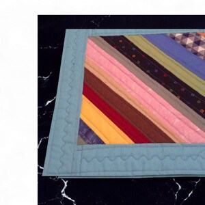 Op de afbeelding: Een handgemaakte, gewatteerde placemat met een lichtblauwe rand. Het midden heeft diagonale strepen in verschillende kleuren, waaronder roze, geel en blauw. De rand heeft een decoratieve, golvende steek.